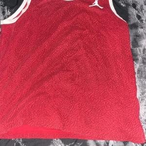Röd Jordan linnetopp med mönster - Säljer en röd linnetopp från Jordan med svart grafiskt mönster över hela tyget. Toppen har vita kantband vid hals och ärmhål samt den klassiska Jumpman-loggan på bröstet. Perfekt för dig som gillar sportig och cool stil.