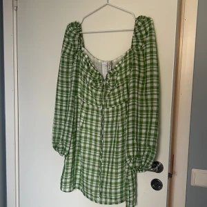 Grön rutig kortklänning från Stradivarius - Superfin grön och vit rutig klänning från Stradivarius i storlek M. Modellen har puffiga långärmar, fyrkantig ringning och dragkedja i ryggen. Tillverkad i 100% bomull, perfekt för dig som gillar en chill och trendig look med lite volym i ärmarna.