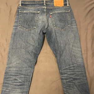 Levi's 512 blå jeans straight fit - Säljer ett par klassiska Levi's 512 jeans i blå tvätt med straight fit. Jeansen har fem fickor, knappgylf och den ikoniska läderpatchen bak i midjan. Snygga slitningar och kontrastsömmar ger en cool vintagekänsla. Perfekt för dig som gillar tidlös stil.