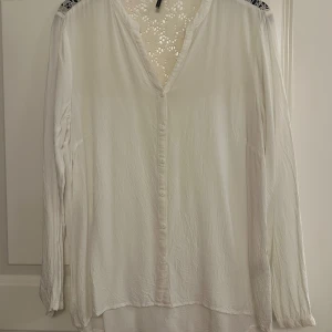 Vit blus med spets från Soyaconcept - Vit långärmad blus från Soyaconcept med v-ringning och knappar framtill. Blusen har snygga spetsdetaljer upptill på axlar och rygg, vilket ger en romantisk vibe. Lätt och flowy passform, perfekt till jeans eller kjol.