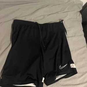 Svarta Nike Dri-Fit shorts M - Svarta sportiga shorts från Nike med vit detalj längst ner och klassisk swoosh-logga på ena benet. Modellen har elastisk midja med snörning och är gjorda i lätt och ventilerande polyester Dri-Fit material. Perfekta för träning eller chill.