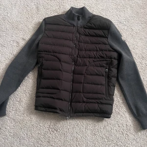 Moncler  cardigan - Snygg svart pufferjacka från Moncler med quiltad front och gråa stickade ärmar. Jackan har hög krage, dragkedja framtill och två fickor med dragkedja. Perfekt för dig som vill ha en stilren och sportig look. Ny Skick  Storlek L men passar M även 