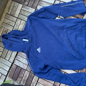 Blå Adidas hoodie med huva - Säljer en marinblå hoodie från Adidas med klassisk logga på bröstet. Tröjan har huva, långa ärmar och en stor magficka framtill. Snygga vita detaljer längs ärmarna ger en sportig vibe. Perfekt för chill eller träning.