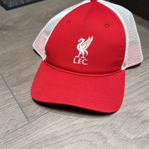 Liverpool FC röd och vit keps - Snygg röd och vit keps med Liverpool FC-logga framtill. Framsidan är röd med broderad vit fågel och L.F.C.-text, medan baksidan har vit mesh och justerbar plastrem. Perfekt för dig som vill visa ditt lag på stan eller matchen.