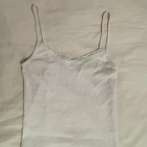 Vit ribbad linnetopp med spets - Säljer en vit ribbad linnetopp med smala axelband och v-ringning dekorerad med spets. Toppen är figurnära och har en enkel, stilren look som passar perfekt till jeans eller kjol. Materialet är mjukt och stretchigt för extra komfort.