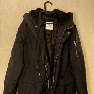 Svart parkajacka från edc, strl S - Svart parkajacka från edc med huva och mysigt foder. Jackan har flera fickor, både med dragkedja och knappar, samt snörning i midjan för snygg passform. Perfekt för kyliga dagar och har en stilren look som funkar till det mesta.