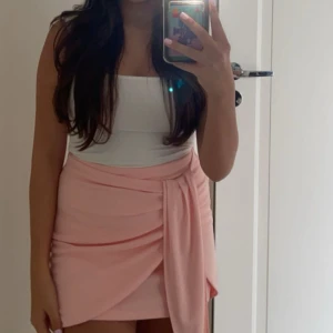 Ljusrosa omlottkjol med volang - Supersöt ljusrosa kortkjol med omlottkänsla och volangdetalj framtill. Kjolen har en draperad look och knytdetalj i sidan som ger en trendig och feminin vibe. Perfekt för dig som gillar en chic och lekfull stil.