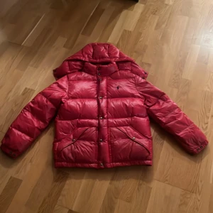 Röd pufferjacka från Polo Ralph Lauren - Säljer en röd pufferjacka från Polo Ralph Lauren med huva och svarta detaljer. Jackan har broderad logga på bröstet, två fickor med knappar och quiltad design. Perfekt för kalla dagar och riktigt snygg streetstil. OBS, en defekt påbaksidan av högra armen (Lagad)