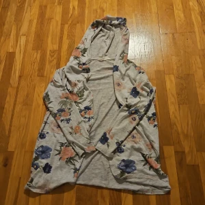 Blommig kofta från H&M med huva - Grå hoodie från H&M med blommigt mönster i blått och rosa. Tröjan har huva och långa ärmar, perfekt för  kyliga sommarkvällar. Den är väldigt mjuk och skön mot huden