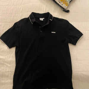 Svart pikétröja från BOSS med vit kant - Klassisk svart pikétröja från BOSS med vit piping längs krage och ärmslut. Tröjan har korta ärmar, knäppning med tre knappar och diskret BOSS-logga på bröstet. Perfekt för en clean och stilren look.