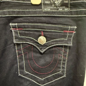 True Religion svarta bootcut jeans - Svarta jeans från True Religion med kontrasterande vita och röda sömmar. Modellen heter Joey Low Rise Flare och har låg midja samt bootcut-ben. Snygga detaljer på fickorna med dekorativa knappar och broderier. Perfekta för dig som gillar en unik jeansstil.