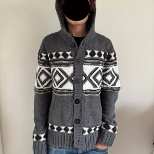 Grå stickad kofta med huva och mönster - Mysig grå stickad kofta med huva och knäppning framtill. Koftan har vita och svarta geometriska mönster och ribbade muddar. Perfekt för kyliga dagar och har praktiska fickor framtill. STORLEK L FITS M