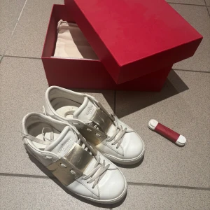 Valentino Garavani vita sneakers med guld - Snygga vita sneakers från Valentino Garavani med bred guldfärgad detalj över snörningen. Skorna är i skinn och har klassisk rund tå, platt sula och vita snören. Kommer med originalkartong och dustbag. Perfekta för dig som vill sticka ut med lyxig touch. Priset kan diskuteras.