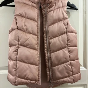 Rosa quiltad dunväst från H&M - Quiltad ljusrosa dunväst från H&M med dragkedja framtill och avtagbar huva med fuskpäls. Västen har en glansig finish och breda, horisontella sömmar som ger en puffig look. Perfekt för lager-på-lager under kyliga dagar.