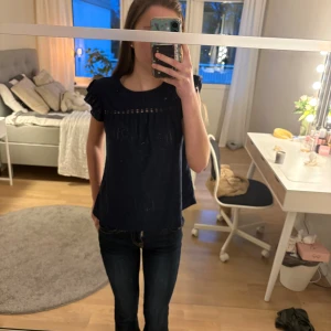 Mörkblå blus med volangärmar - Säljer en mörkblå blus med korta volangärmar och dekorativt hålmönster upptill. Blusen har en lös passform. Passar till både jeans och kjol.