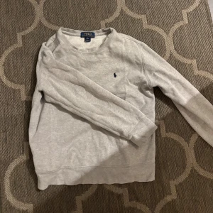 Sweatshirt Polo Ralph Lauren (10-12) - En stilren ljusgrå sweatshirt från Polo Ralph Lauren med klassisk rund hals och broderad marinblå logga på bröstet. Tröjan är i mjuk bomull och har långa ärmar, perfekt för en avslappnad look. Storlek M i barnstorlek 10-12 år.