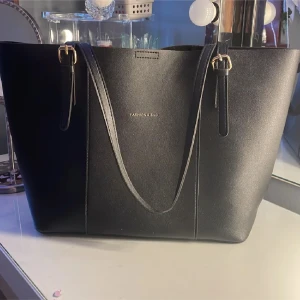 Svart axelväska från Fashion & Bag - Stilren svart axelväska med slät yta och guldfärgade detaljer. Väskan har två långa axelremmar med spännen och rymligt innerfack som stängs med magnetknapp. Perfekt för laptop och allt du behöver under dagen.