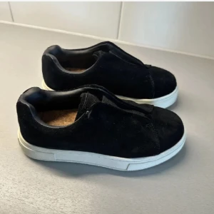 Svarta Eytys - Svarta sneakers i mjuk mocka med vit platt sula. Skorna har en stilren och enkel design utan synlig snörning, vilket ger en clean look. Perfekta för dig som gillar minimalistisk stil och vill ha något som funkar till det mesta.