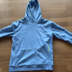 Ljusblå hoodie från Scotch & Soda - Ljusblå hoodie från Scotch & Soda med huva och magficka. Diskret limegrön textdetalj vid fickan och patch bak nedtill. Perfekt för en chill och avslappnad stil. Mjukt material som känns skönt mot huden.