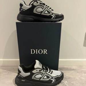 Svarta och silver Dior sneakers - Snygga Dior sneakers i svart och silver med tydlig CD-logga på sidan. Skorna har en chunky platt sula, rund tå och snörning. Materialet är en mix av syntet och mesh som ger en modern och sportig vibe. Perfekta för dig som gillar exklusiva märken och streetstyle.