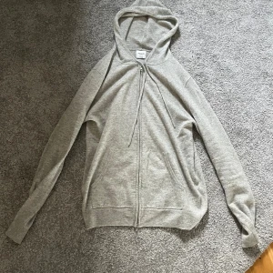 Kashmir hoodie från John Henric - Säljer en stilren grå hoodie från John Henric med dragkedja framtill och huva med snörning. Tröjan har långa ärmar, ribbade muddar och två fickor framtill. Perfekt för en avslappnad look.