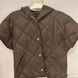 Brun quiltad pufferväst med huva - Quiltad brun pufferjacka med korta ärmar och huva. Jackan har tryckknappar framtill och två fickor. snyggt rutmönster i materialet. Perfekt för lager-på-lager och chill höst- eller vårstil.