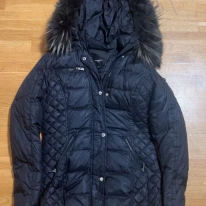 Svart Rockandblue dunjacka med päls - Snygg svart dunjacka från Rockandblue med stor fluffig fuskpäls på huvan. Jackan har quiltade partier, flera dragkedjor och en badge på ärmen. Perfekt för kalla dagar och har en riktigt schysst streetstil.