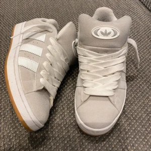 Adidas Campus sneakers i ljusgrå mocka - Adidas Campus sneakers i ljusgrå mocka med klassiska vita ränder på sidorna och gummisula i beige. Skorna har rund tå, platt sula och snörning. Adidas logga på plösen och hälen. Perfekt för dig som gillar retrostil och streetwear.