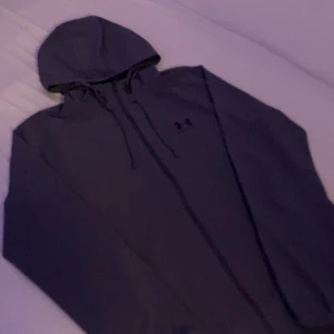 Grå vindjacka från Under Armour - Säljer en grå vindjacka från Under Armour Storm med huva och dragsko. Jackan är tillverkad i lätt syntetmaterial och har en stilren look med diskret logga på bröstet. Perfekt för blåsiga dagar och enkel att matcha med det mesta.