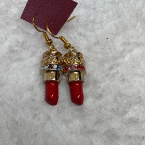 Örhängen med läppstift och strass - Unika örhängen formade som röda läppstift med guldfärgade detaljer och glittrande strass i olika färger runt toppen. Perfekt för dig som vill sticka ut och addera lite färg och glamour till din stil.