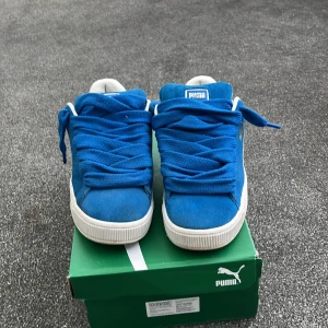 Blå Puma Suede sneakers strl 41 - Snygga blå Puma Suede sneakers med breda blå skosnören och vit sula. Skorna har klassisk låg profil, vit sidodetalj och guldfärgad Puma-logga på sidan. Ovandelen är i mocka och insidan är vit med blå detaljer. Perfekta för dig som gillar retrostil. Låda och ett till vitt skosnöre igår också.