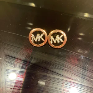Roséguldiga MK örhängen med strass - Snygga örhängen från Michael Kors i rund form med MK-logga i mitten. Örhängena är guldiga och dekorerade med glittrande strass på loggan, vilket ger en lyxig känsla. Perfekt för dig som gillar statement-smycken och vill ha något som syns.