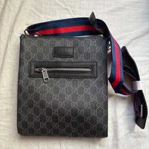 Svart axelväsk med GG-mönster - Snygg svart axelväska med GG-mönster över hela väskan. Väskan har en bred axelrem i blått och rött, samt en praktisk dragkedja framtill. perfekt för dig som gillar designer och streetstyle.