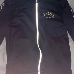Svart zip hoodie från Hugo Boss - Svart zip hoodie från Hugo Boss med dragkedja framtill och guldiga logodetaljer på bröstet och dekorativa ränder på ärmarna. Perfekt för en stilren och sportig look. Den är i storlek S.