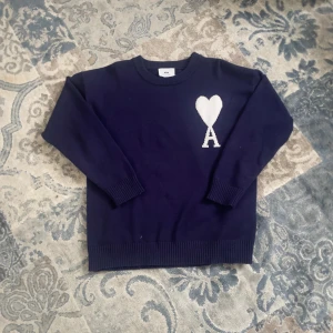 Mörkblå stickad tröja från Ami - Säljer en mörkblå stickad tröja från Ami Alexandre Mattiussi med vit hjärtlogga och A på bröstet. Tröjan har rund hals, ribbade muddar och är långärmad. Perfekt för dig som gillar stilrena och trendiga plagg.