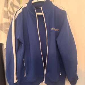 Blå Palm Angels ziptröja med vita ränder - Säljer en blå ziptröja från Palm Angels med vita ränder längs ärmarna och dragkedja framtill. Tröjan har hög krage, två fickor med dragkedja och logga på bröstet. Perfekt för en sportig och trendig look. Priset går diskuteras elr trade