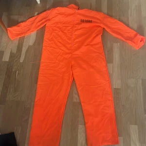 Orange fängelsedräkt med dragkedja - Säljer en helkropps orange fängelsedräkt med svarta siffror tryckta på bröstet. Dräkten har lång ärm, rak passform och dragkedja framtill. Perfekt för temafest eller maskerad. Materialet känns som polyester och färgen är riktigt stark orange.