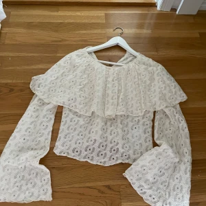 Vit spetsblus med volang - Superfin vit blus i spets med stora volanger över axlarna och vida långa ärmar. Blusen har ett romantiskt mönster och är perfekt för dig som gillar en söt och trendig stil. Den är lätt och luftig, perfekt för vår och sommar.