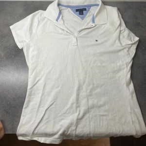 Vit piké från Tommy Hilfiger - Vit pikétröja från Tommy Hilfiger med klassisk krage och korta ärmar. Tröjan har blå detaljer på insidan av kragen och en liten broderad logga på bröstet. Tillverkad i mjuk bomull som är skön mot huden. Perfekt för en clean och stilren look.