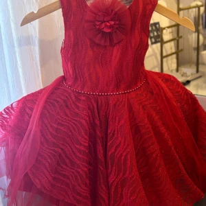 Röd festklänning från Newe Baby - Supersöt röd kortklänning från Newe Baby med glittrigt vågmönster, voluminös kjol i flera lager tyll och pärldetalj i midjan. Framtill sitter en stor blomma i tyll som extra detalj. Klänningen är ärmlös och har dragkedja bak.