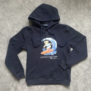 Mörkblå Polo Bear hoodie från Ralph Lauren - Säljer en mörkblå hoodie från Ralph Lauren med Polo Bear-tryck där björnen surfar på en våg. Tröjan har huva med snörning, magficka och långa ärmar. Skön och stilren, perfekt för dig som gillar streetwear och klassiska motiv.