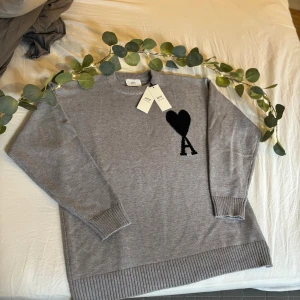 Grå stickad tröja från Ami Paris - Snygg grå stickad tröja från Ami Paris med svart hjärta och A-logga på bröstet. Tröjan har rund hals, ribbade muddar och är tillverkad i mjuk ull. Perfekt för dig som gillar stilrena och trendiga plagg med en twist.