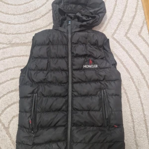 Svart dunväst från Moncler med huva - Svart dunväst från Moncler med avtagbar huva och två praktiska fickor med dragkedja. Västen har quiltad design och Moncler-logga broderad på bröstet. Perfekt för lager-på-lager och snygg till streetwear. Stängs med dragkedja framtill.