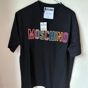 Svart Moschino t-shirt med färgglatt tryck - Cool svart t-shirt från Moschino med stort, broderat MOSCHINO-tryck i flera färger på bröstet. Klassisk rund hals och korta ärmar. Materialet är mjuk bomull som känns skönt mot huden. Perfekt för dig som gillar statement-plagg och vill sticka ut.