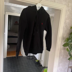 Svart hoodie från Polo Ralph Lauren - Svart hoodie från Polo Ralph Lauren med klassisk röd logobrodyr på bröstet. Tröjan har huva med snörning, stor magficka och långa ärmar. Tillverkad i mjuk bomull som är skön att ha på sig. Perfekt för chill dagar och streetwear-stil.Helt ny