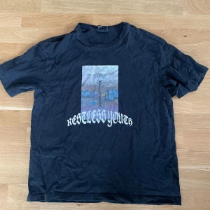 Svart t-shirt Restless Youth - Svart t-shirt från Weekday med tryck framtill som visar en basketkorg och graffiti, samt texten 'Restless Youth' i vit gotisk stil. Klassisk rund hals och korta ärmar. Materialet är mjuk bomull, perfekt för en chill streetwear-look.