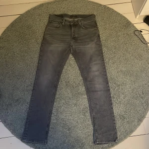 Grim Tim Nudie Jeans W30 L30 - Säljer ett par grå skinny jeans från Nudie Jeans i modellen ”grim tim” i storleken W30 L30 med klassisk femficksdesign och svarta detaljer på bakfickorna. Jeansen har en smal passform och är tillverkade i stretchig denim för extra komfort. Perfekta för dig som gillar en stilren och modern look.