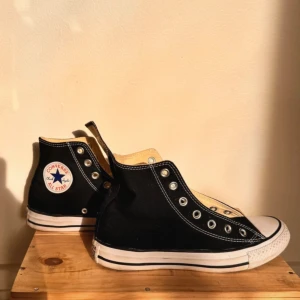 Svarta Converse All Star High 37 - Klassiska svarta Converse All Star med vit tå och sula. Skorna har rund tå, platt gummisula och snörning. Ovandelen är i slitstark bomullscanvas med vita kontrastsömmar och Converse-logga på sidan. Perfekta för en avslappnad streetstil. Storlek 37
