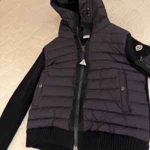  Moncler CARDIGAN - Snygg svart pufferjacka från Moncler med stickade ärmar och quiltad framsida. Jackan har huva med snörning, dragkedja framtill och två fickor med tryckknappar. Ärmarna är ribbstickade och har en Moncler-logga på vänster arm. 10/10