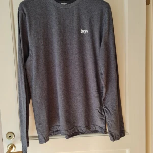 Grå långärmad tröja från DKNY strl XL - Snygg grå långärmad tröja från DKNY i storlek XL. Tröjan har en enkel och stilren design med liten vit DKNY-logga på bröstet. Tillverkad i 100% bomull. Aldrig använd.